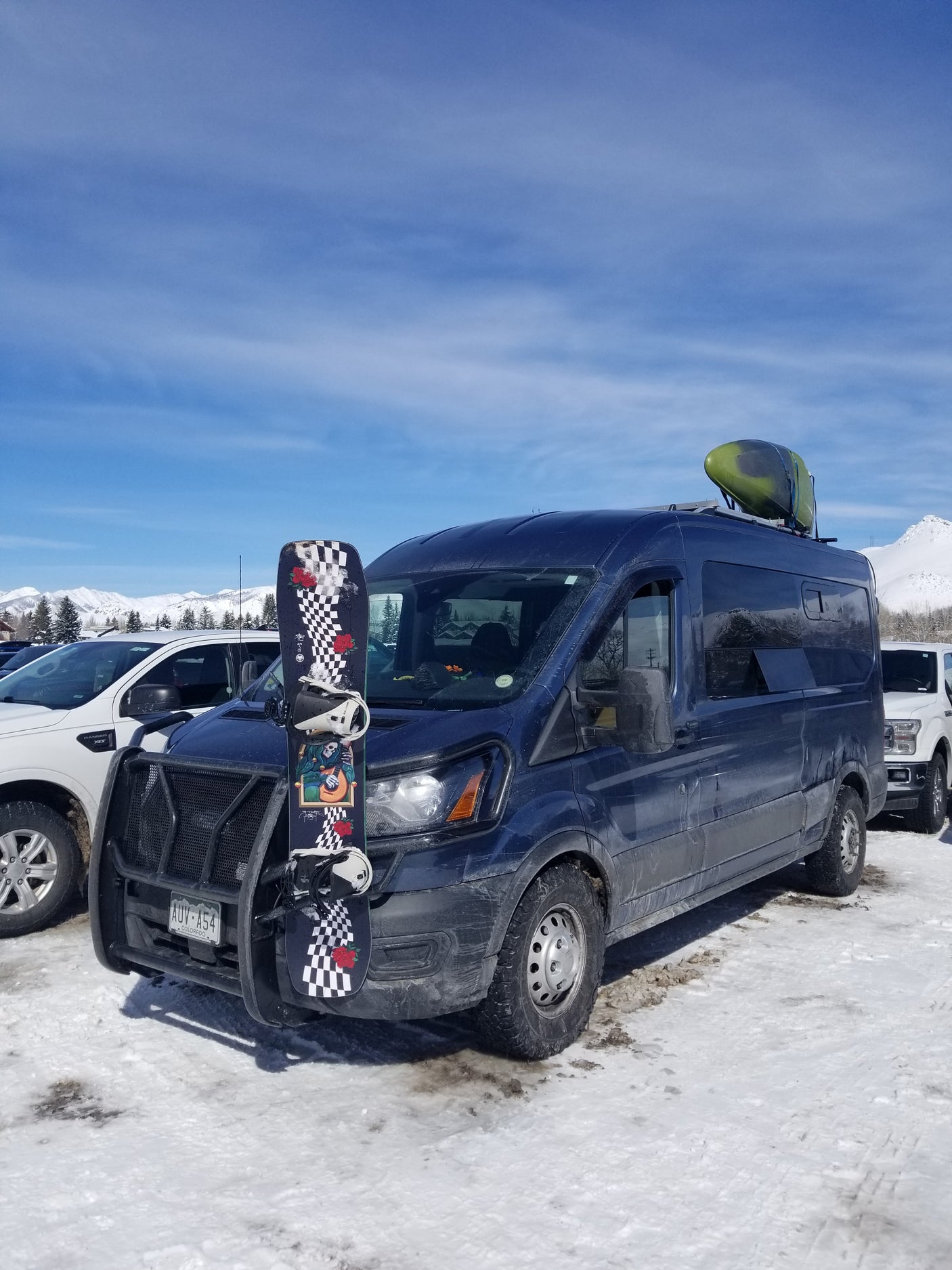 An adventure-ready RV rental van, fully loaded with a snowboard and kayak, parked in a snowy mountain setting.