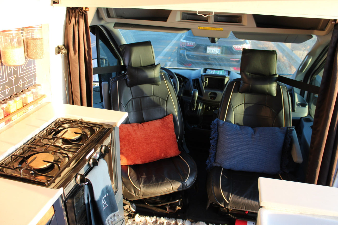 RV rental interior: Front cabin with black seats, dashboard view, and a built-in kitchen with a stove on the left.