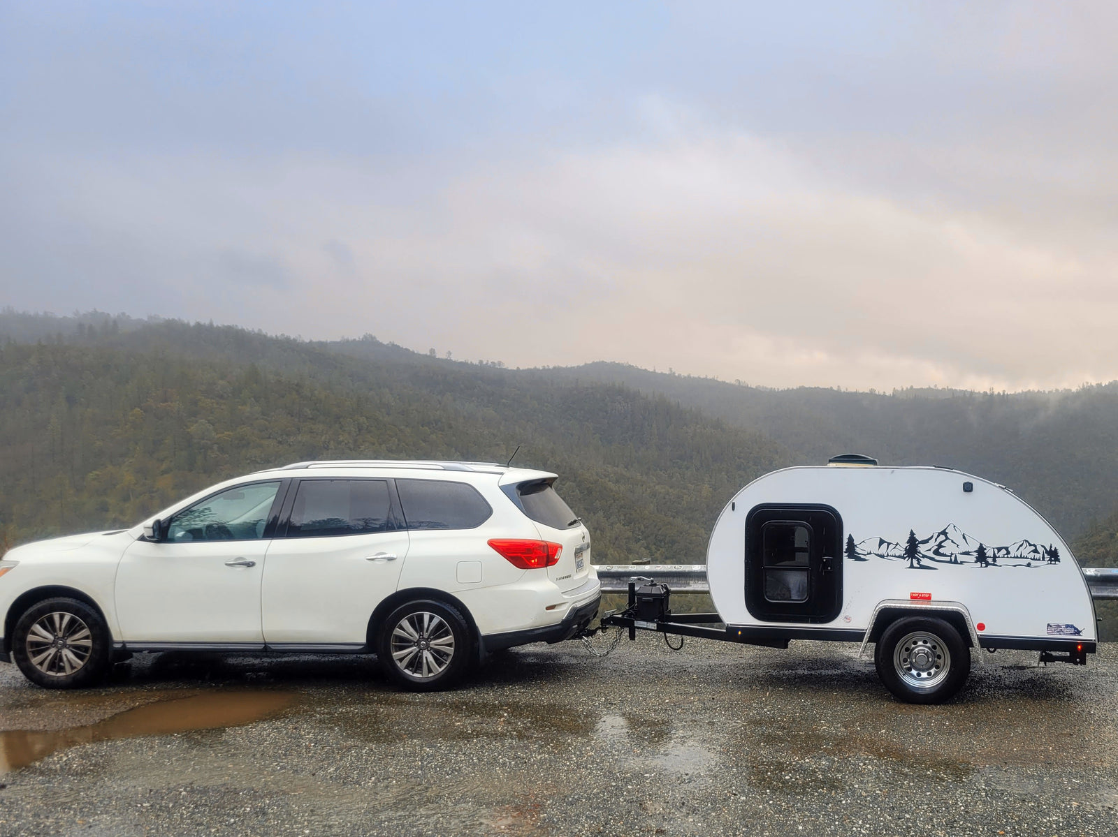RV rental: White SUV towing a teardrop travel trailer on a wet mountain road, ready for a camping road trip.
