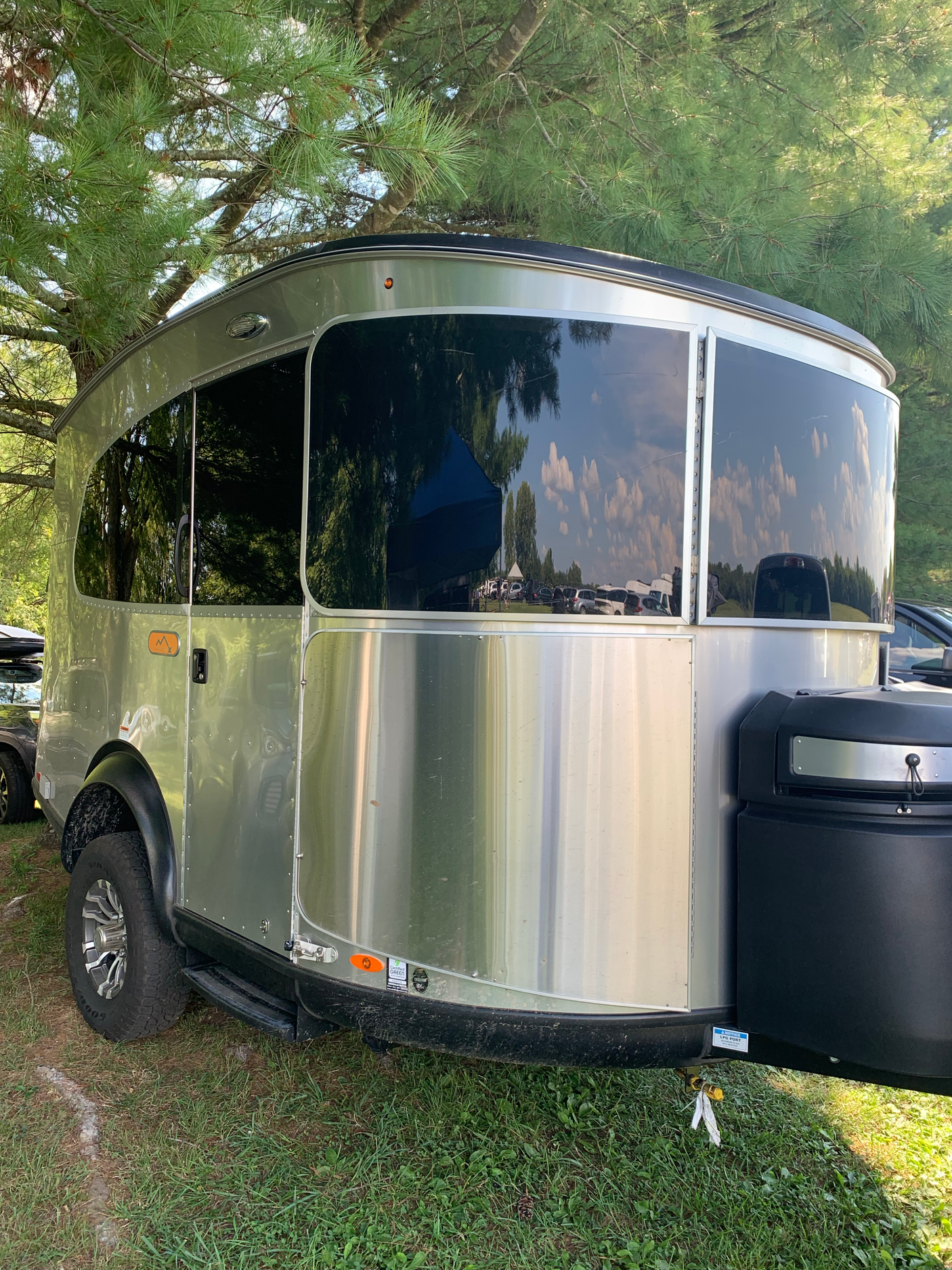 Sleek silver RV rental travel trailer parked under pine trees. Its large reflective windows show sky and cars, perfect for outdoor adventures.