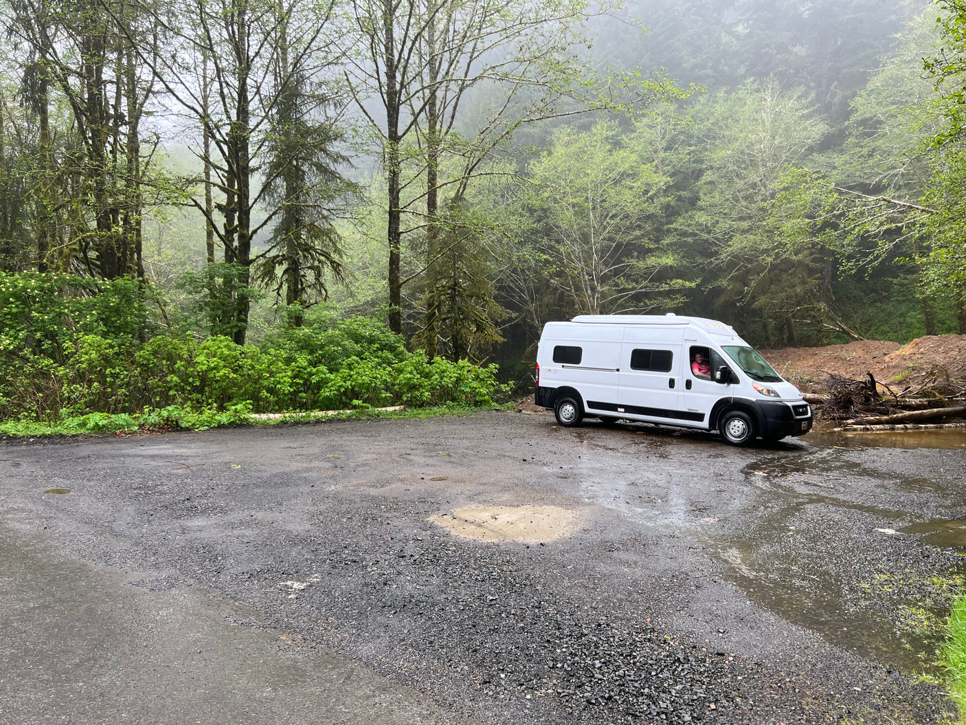 White rv rental parked on a wet gravel lot, surrounded by a misty forest. A person smiles from the driver's seat.