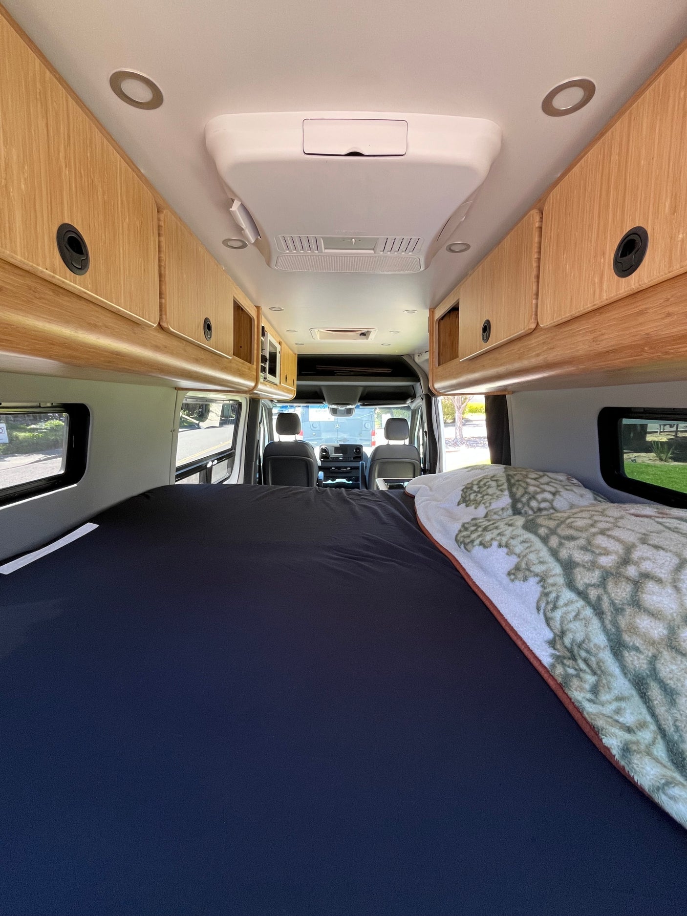Spacious RV rental interior featuring a comfortable bed, wood overhead cabinets, AC unit, and driver's cabin view.