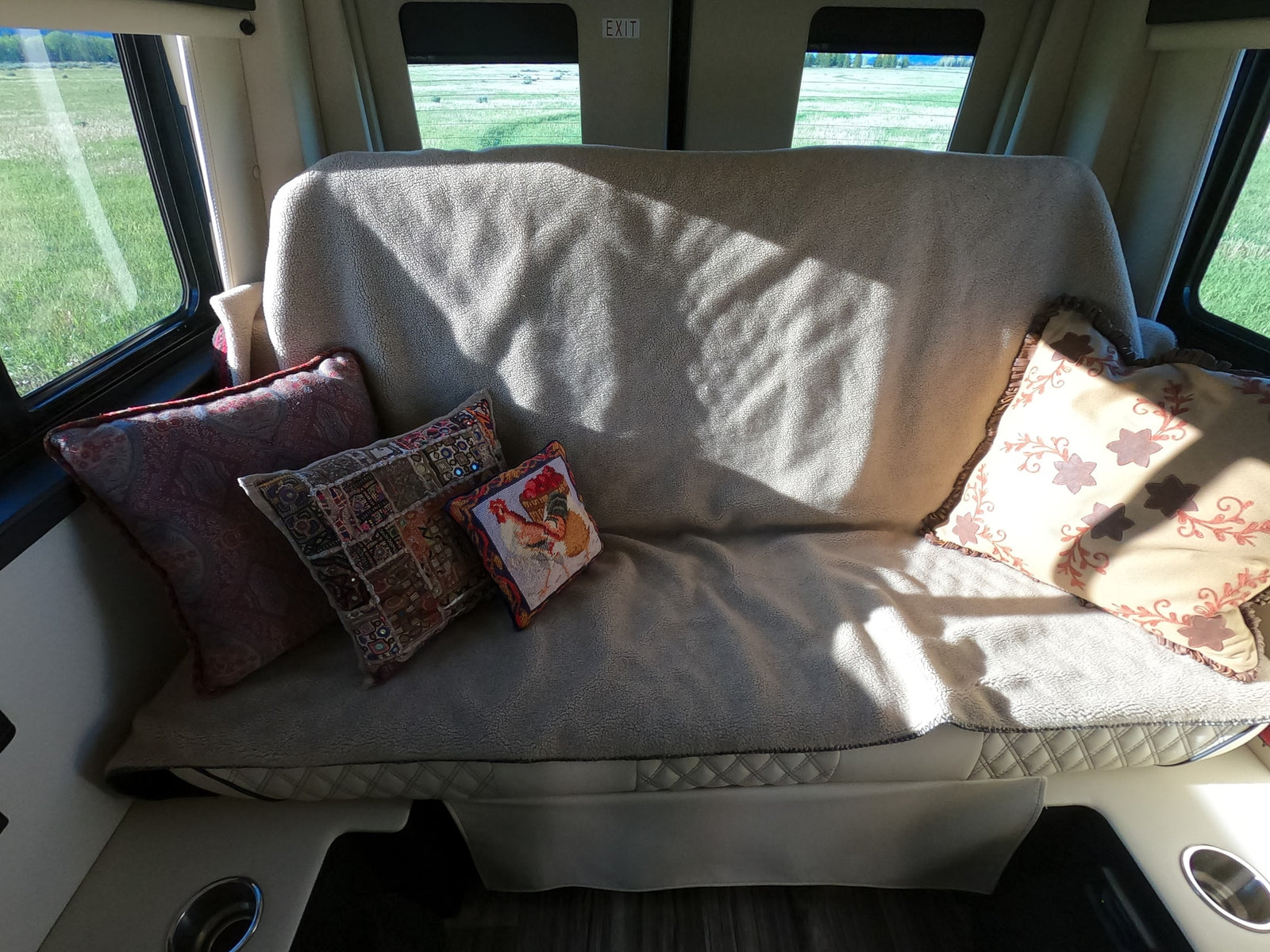 Cozy RV rental interior featuring a sofa with decorative pillows, sunlight, and a scenic view of green fields through the windows.