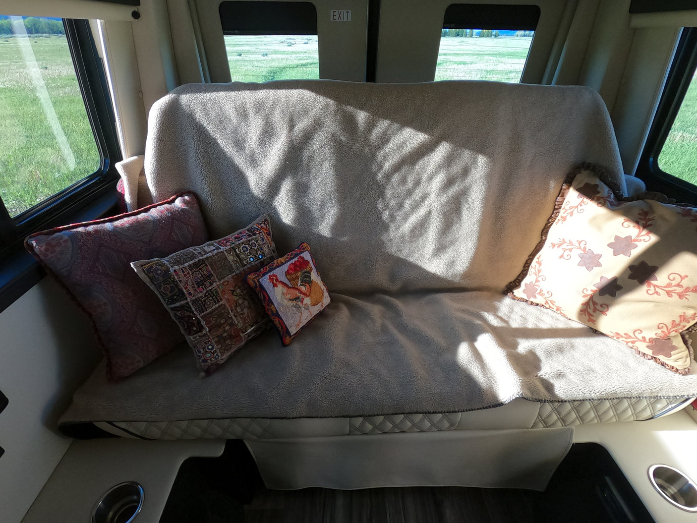 Cozy RV rental interior featuring a sofa with decorative pillows, sunlight, and a scenic view of green fields through the windows.