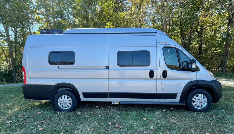 Side view of a silver Class B RV rental camper van, parked on grass in front of green trees.