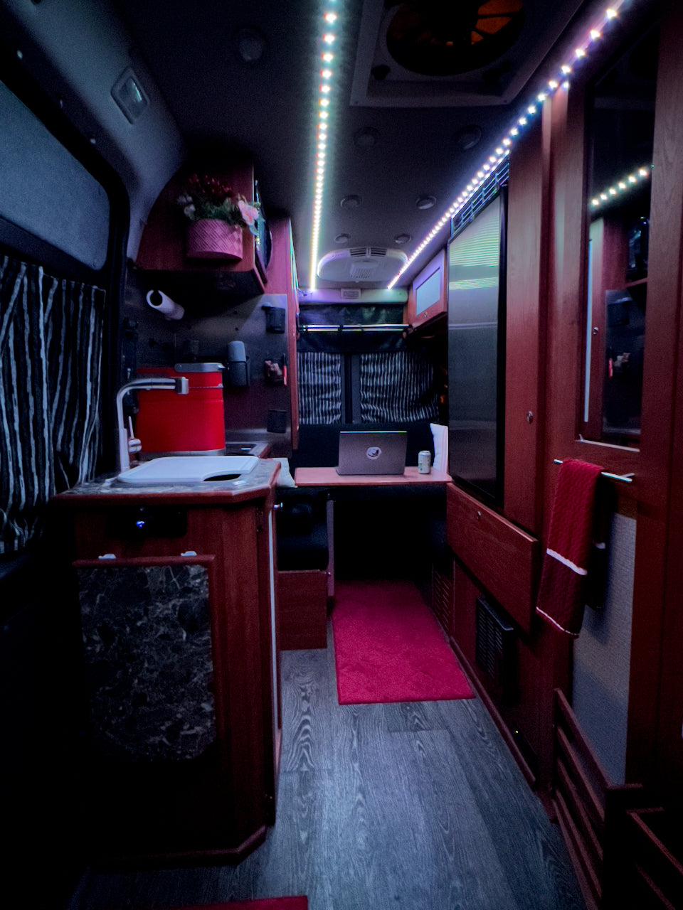 Cozy RV rental interior features dark wood cabinetry, a compact kitchen, workspace, & bright LED lighting.