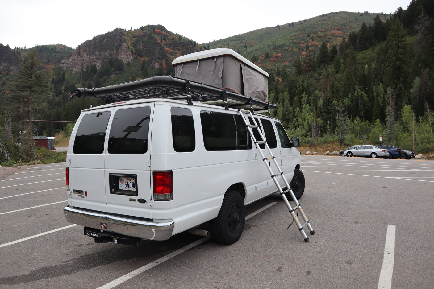 Explore nature with this white rv rental camper van, featuring a rooftop tent & ladder, parked by autumn mountains.