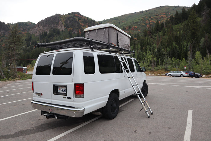 Explore nature with this white rv rental camper van, featuring a rooftop tent & ladder, parked by autumn mountains.