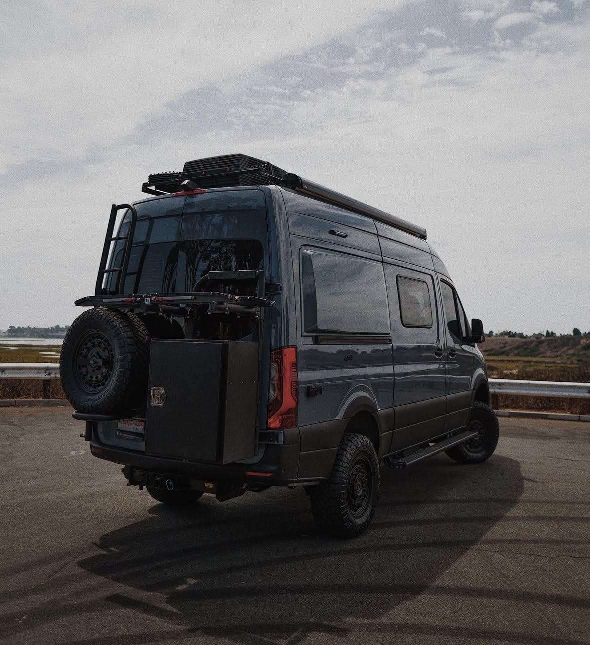 Dark gray Mercedes Sprinter van, an ideal rv rental. Features off-road tires, a ladder, storage box, and roof rack. — photo 3