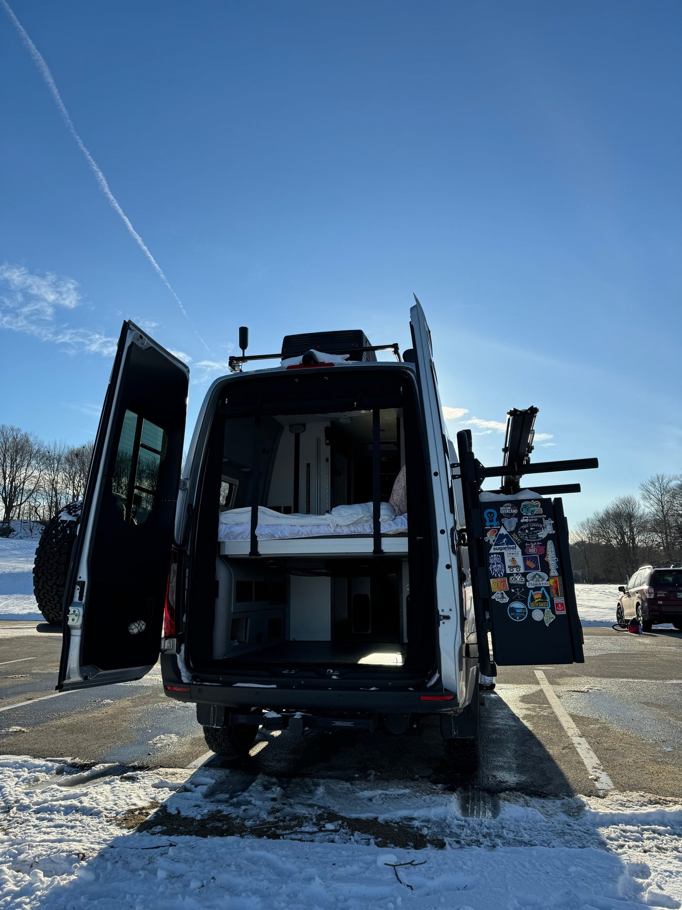 Rear of an RV rental with open doors showcasing a camper bed & gear space. Sticker-covered rack & spare tire ready for adventures!