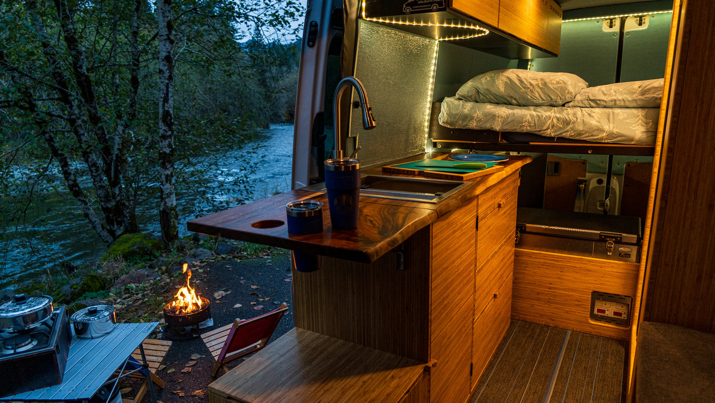 Cozy RV rental interior with kitchen, bed, and outdoor view of a river and campfire at dusk. Perfect for adventurers.