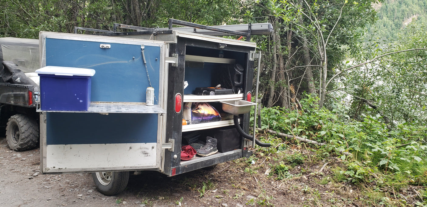 Custom trailer's outdoor kitchen (sink, shelves) for RV rental camping, amidst lush trees by a body of water.