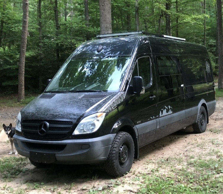 parked by a rock cliff.|Explore the woods in this black Mercedes Sprinter camper van with off-road tires. Your next RV rental adventure awaits