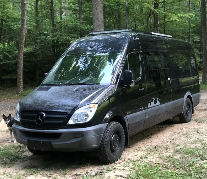 parked by a rock cliff.|Explore the woods in this black Mercedes Sprinter camper van with off-road tires. Your next RV rental adventure awaits