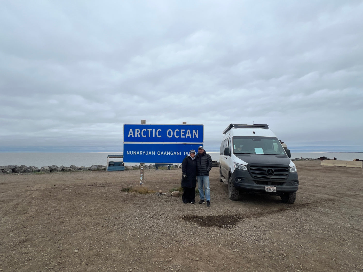 RV rental adventure to the Arctic Ocean! Two people pose by the sign with their van, under a vast cloudy sky.