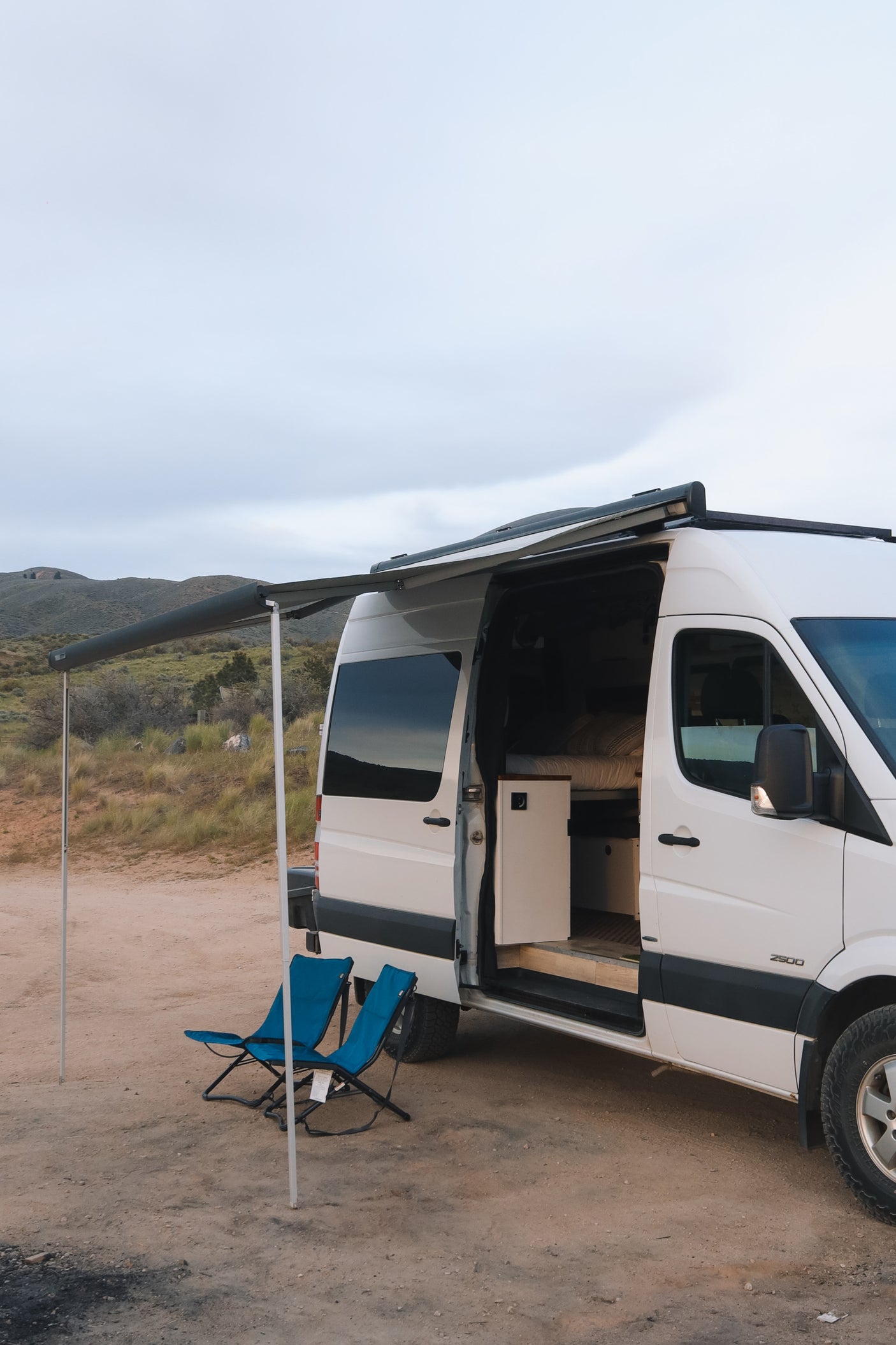 RV rental camper van with open side door and extended awning, providing shade for two blue chairs in a rustic outdoor setting.