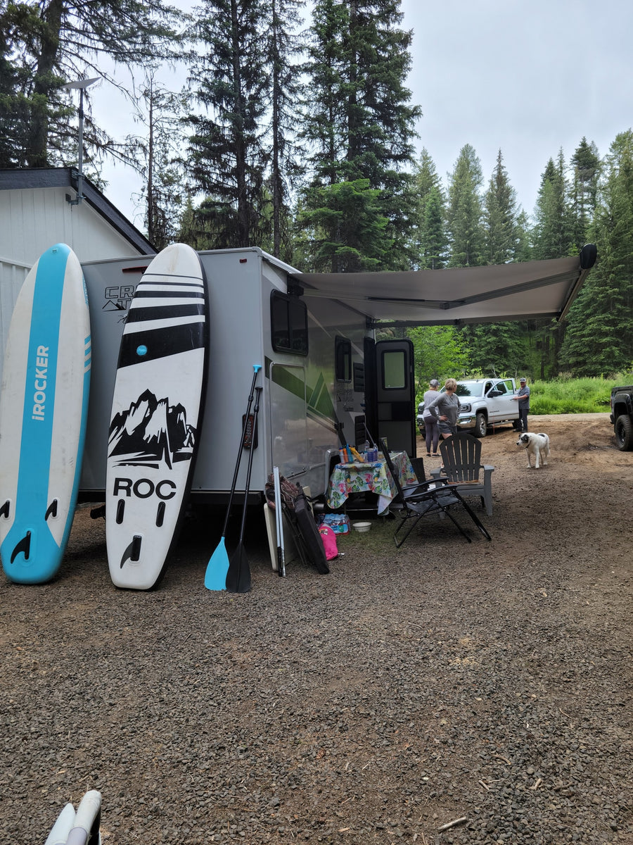 An RV rental parked in a wooded camp, featuring paddleboards, people, and a dog enjoying the outdoors.
