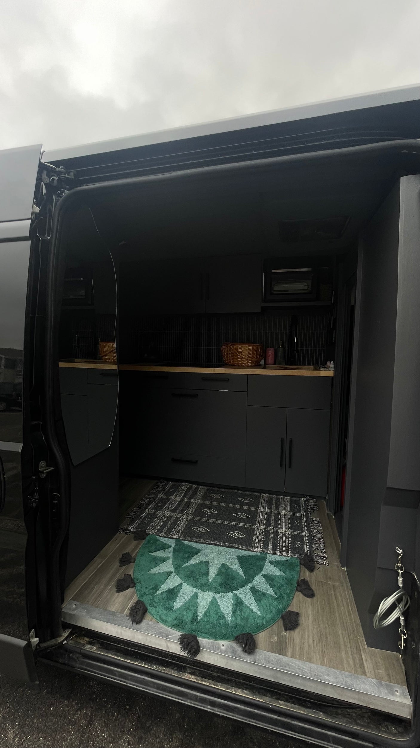 Modern RV rental interior with a compact dark kitchen, wood counters, and patterned rugs seen from the open side door.