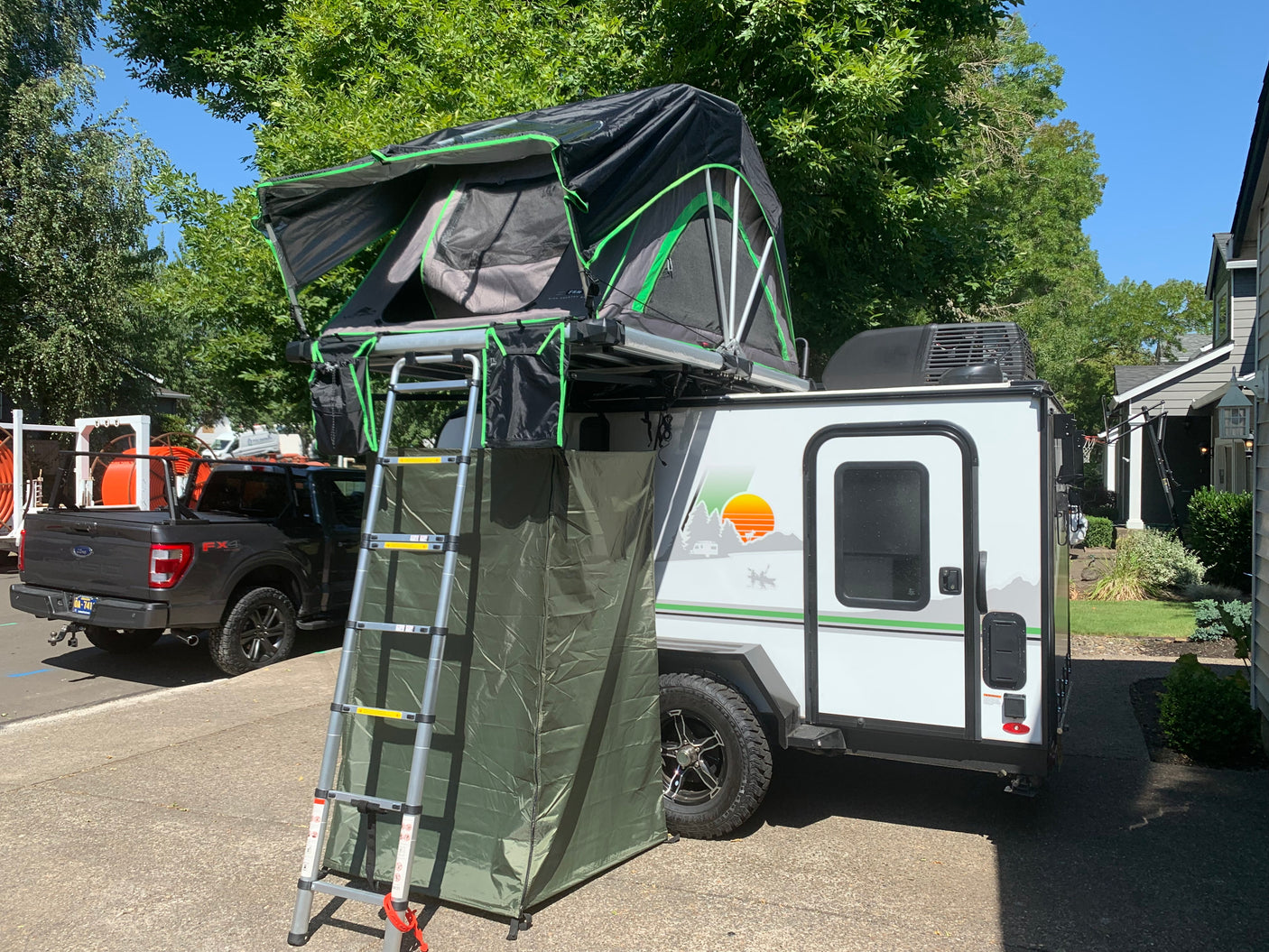 This RV rental, a small travel trailer, features a roof-top tent with ladder & privacy enclosure, parked roadside.