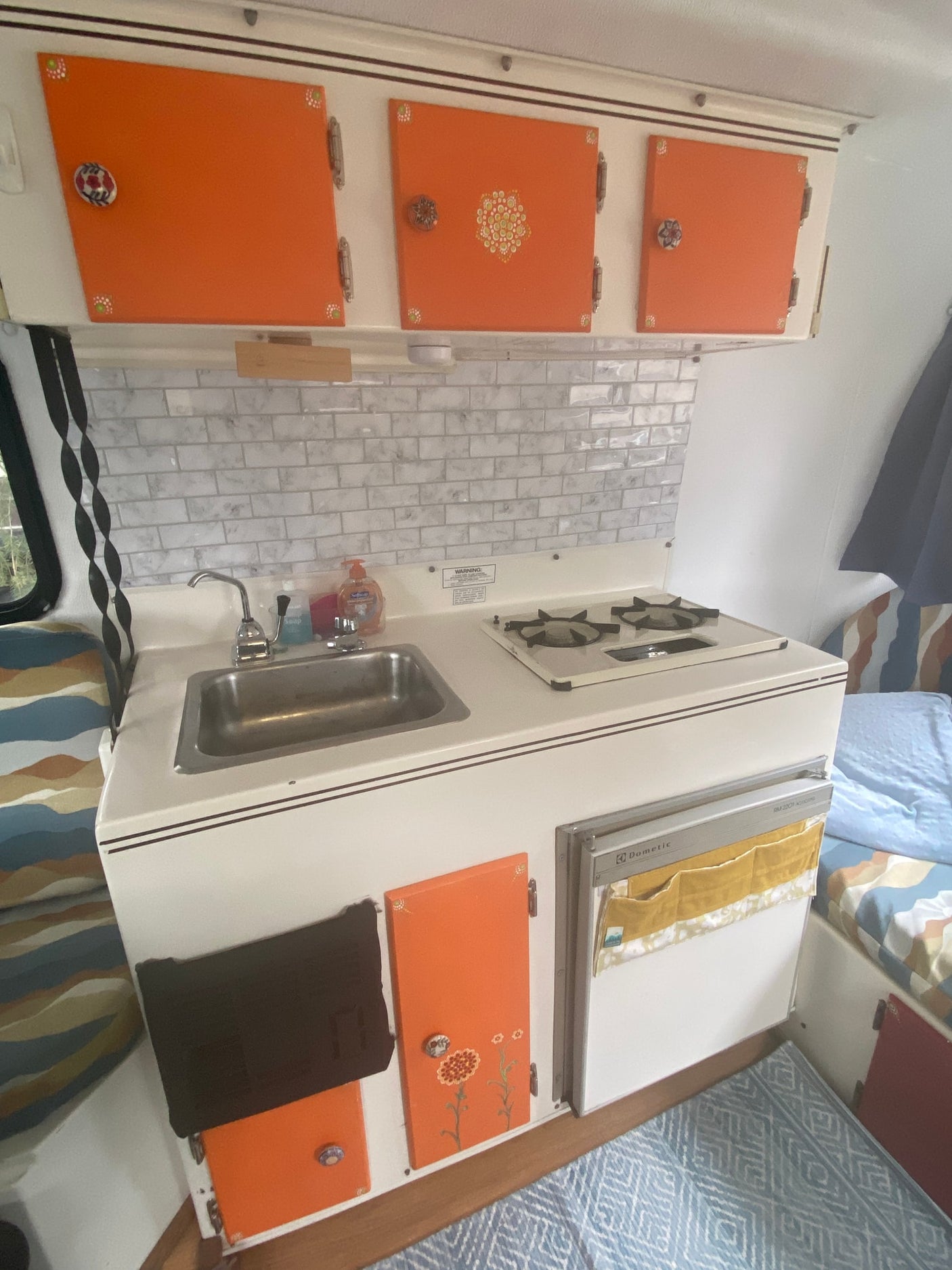 Bright orange cabinets and white appliances in a retro-themed RV rental kitchen, with a sink, stove, and tile backsplash.