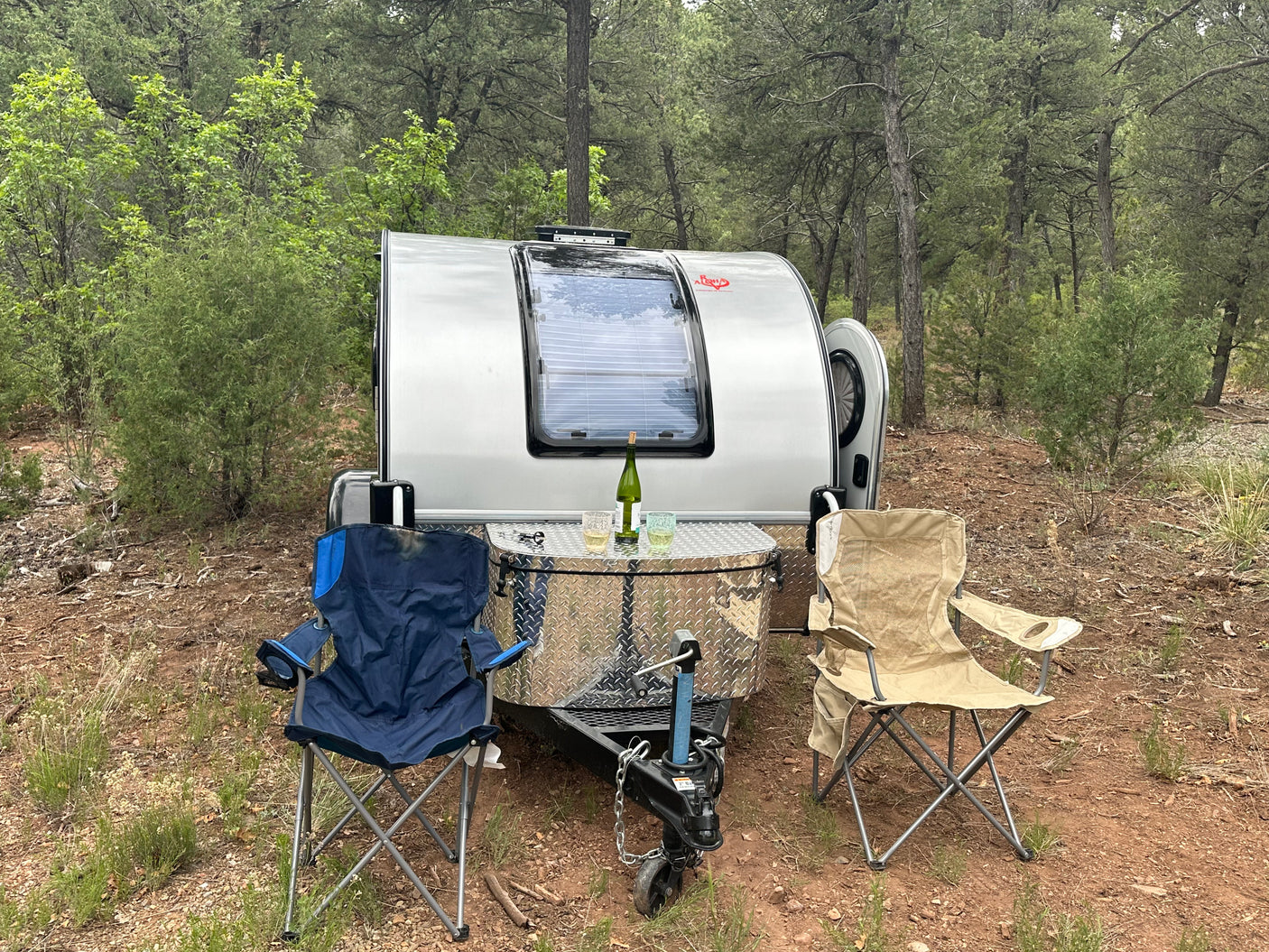 Cozy RV rental setup: a silver teardrop trailer, two chairs, and wine at a wooded campsite. Perfect outdoor escape.