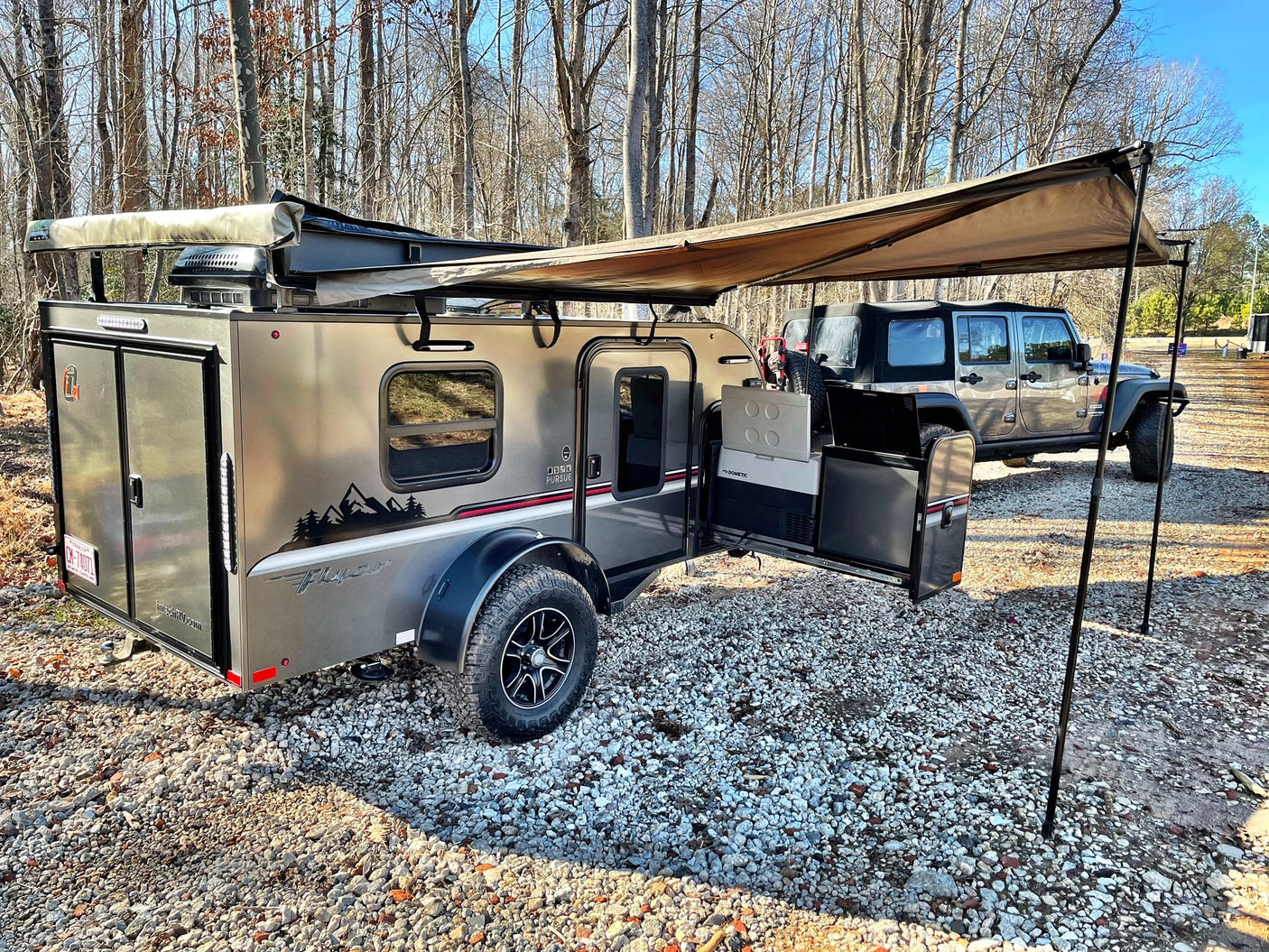 Adventure-ready RV rental: A modern teardrop trailer with an extended awning and Jeep on a gravel campsite outdoors.