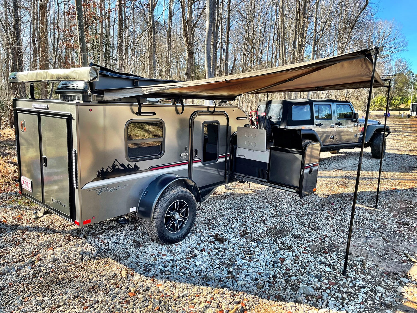 Adventure-ready RV rental: A modern teardrop trailer with an extended awning and Jeep on a gravel campsite outdoors.