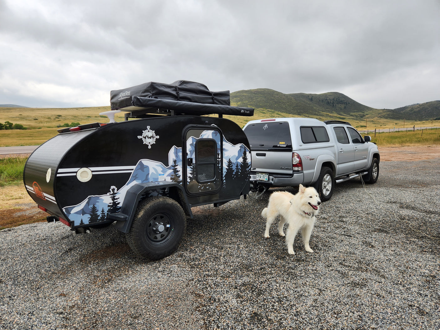 Exploring with an RV rental: a black teardrop trailer, silver truck, & white dog on a gravel lot, backed by cloudy hills.