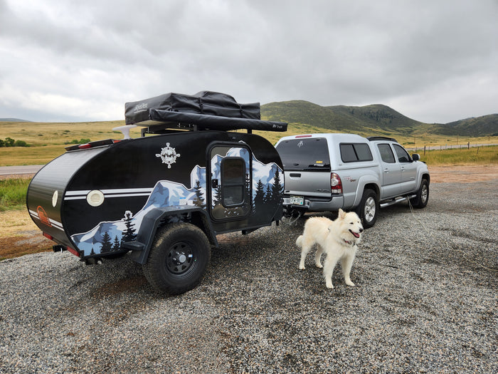 Exploring with an RV rental: a black teardrop trailer, silver truck, & white dog on a gravel lot, backed by cloudy hills.