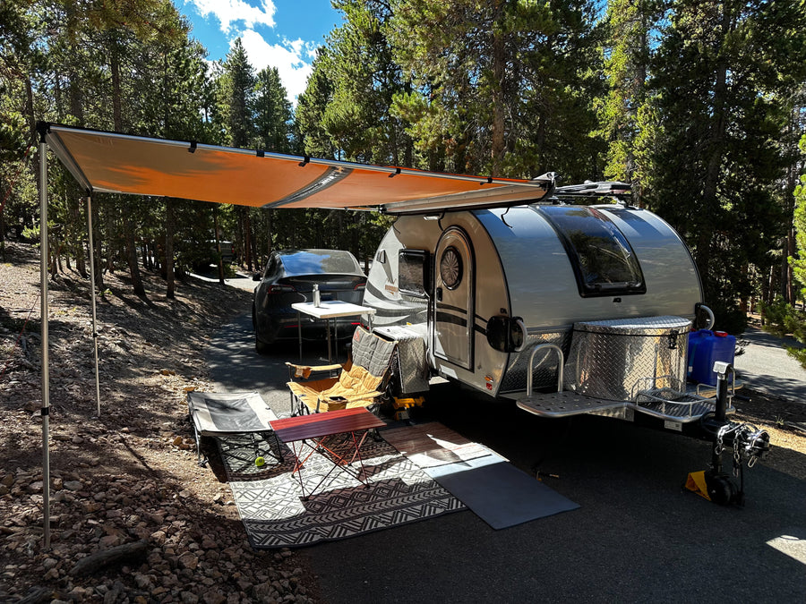 Sunny forest RV rental setup: teardrop trailer with awning, chairs, and Tesla parked nearby.