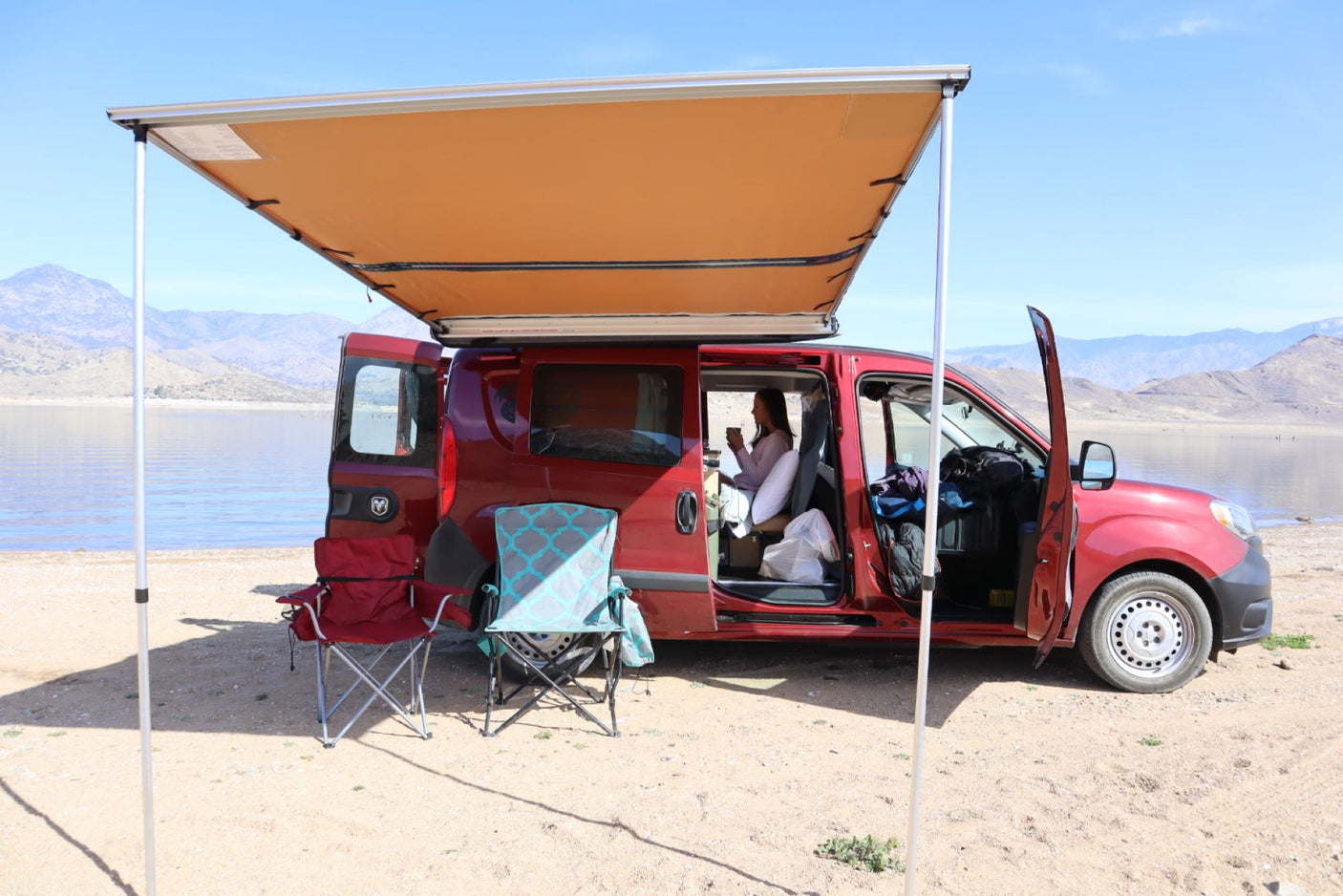 looking out at a lake & mountains. A perfect RV rental escape!|Lakeside camping in a red camper van with an awning. A perfect RV rental for scenic outdoor adventures.|Relax in this cozy rv rental van setup! Features a bed & fridge for comfortable camping and outdoor adventures.|RV rental kitchen setup: a pot