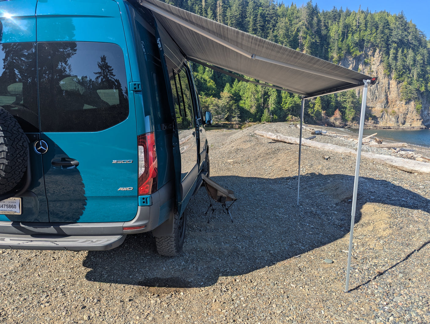 Teal Sprinter RV rental with extended awning on a sunny rocky beach, ready for coastal outdoor adventures.