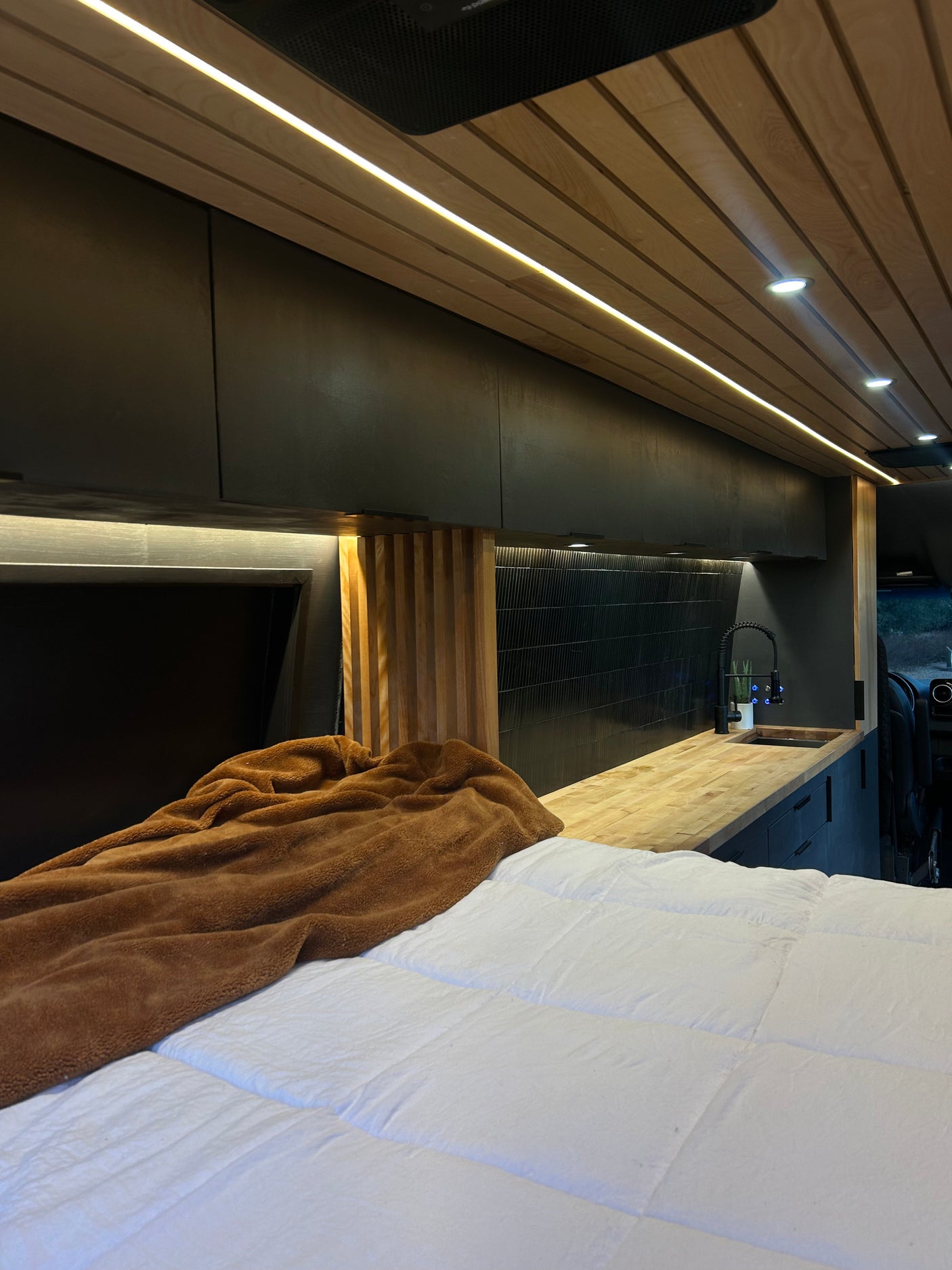 Inside a stylish RV rental: cozy bed, elegant wood-slat ceiling lights, and fully equipped kitchenette.