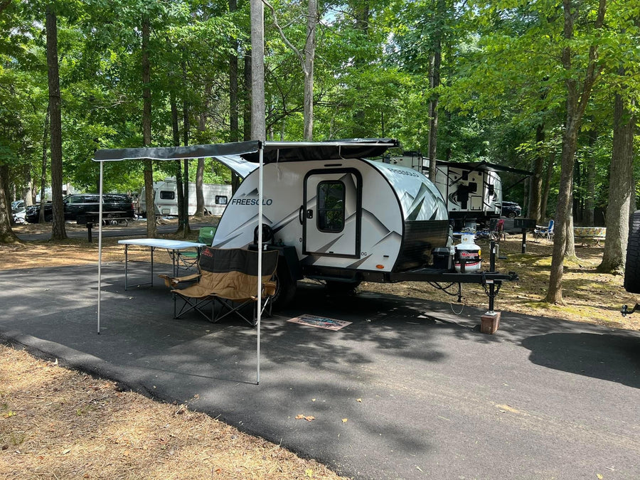 Enjoy your RV rental experience! A white teardrop trailer is set up at a shady campground site with an awning, table, and chairs.