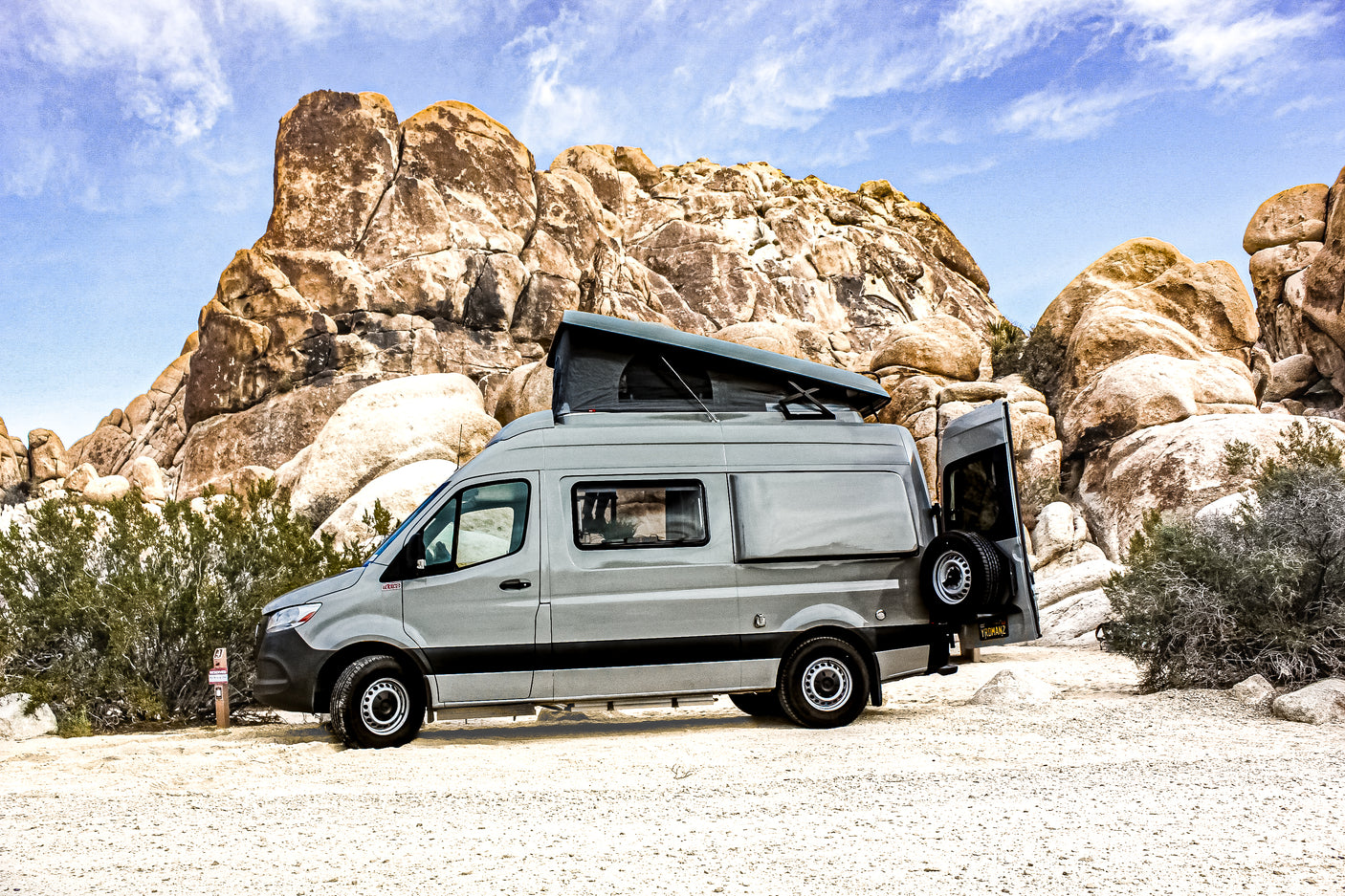 Explore with this grey RV rental camper van, featuring a pop-top tent, parked among desert rocks. Ideal for outdoor adventures.
