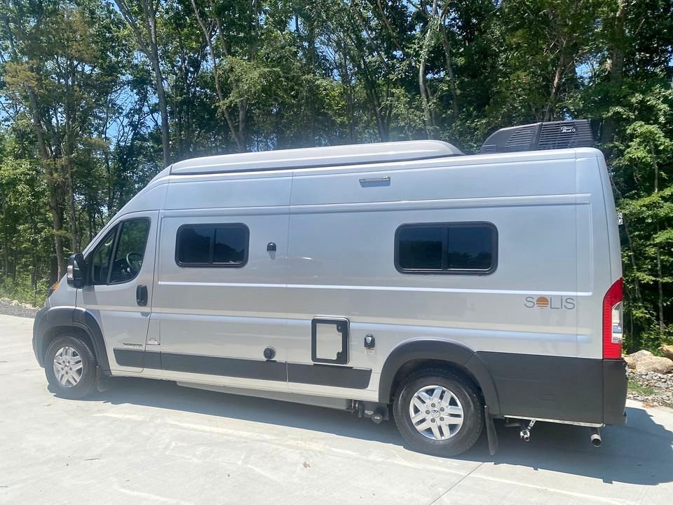 Silver Winnebago Solis camper van, ideal for rv rental adventures. Parked on concrete with lush green trees in background.
