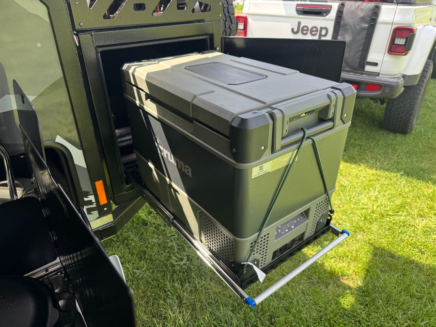 A heavy-duty Truma portable fridge, secured with a bungee, slides out of an RV rental unit on green grass.
