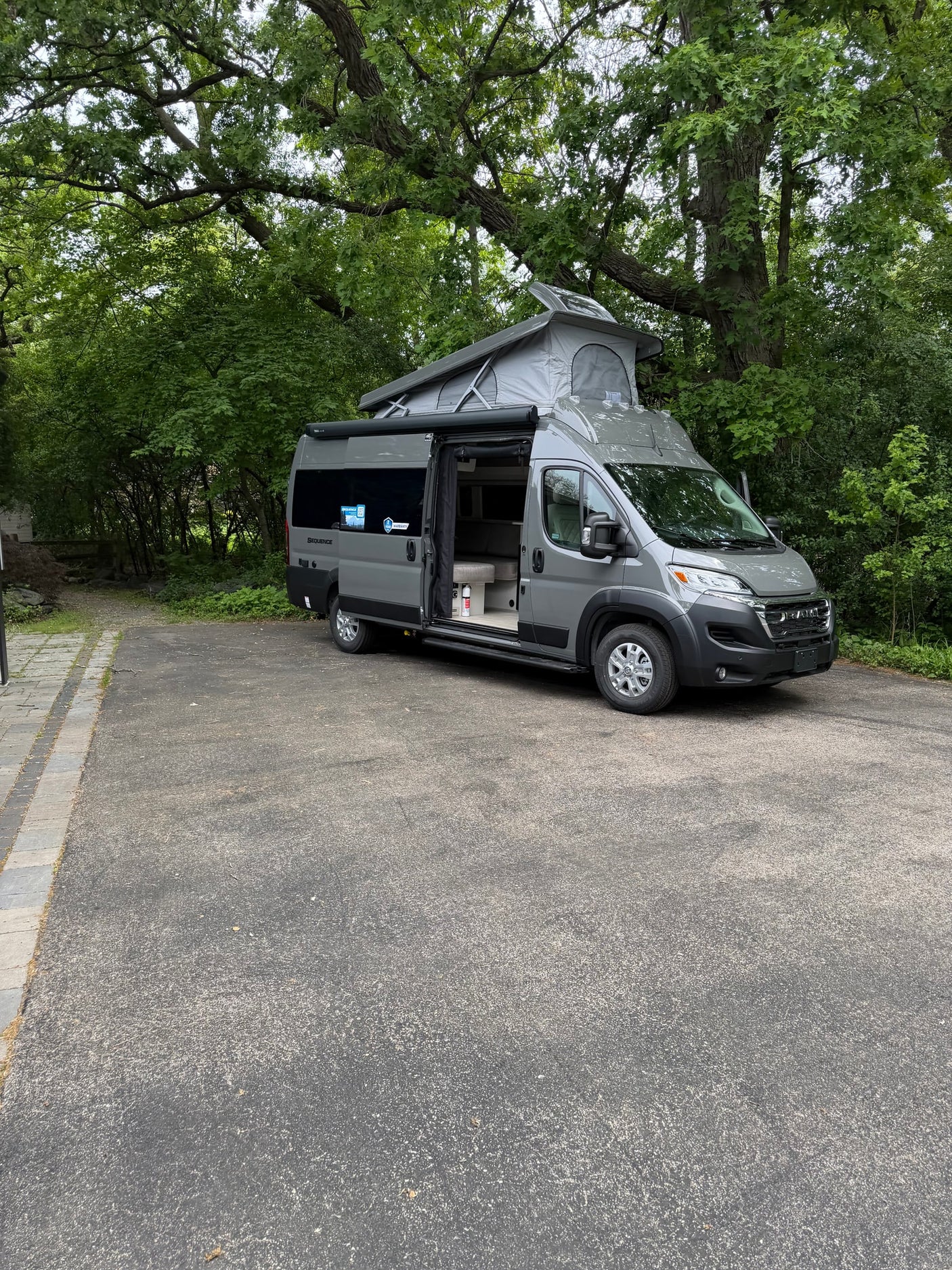 A modern gray camper van with an extended pop-top roof is parked on asphalt, ready for your rv rental adventure amidst lush trees.