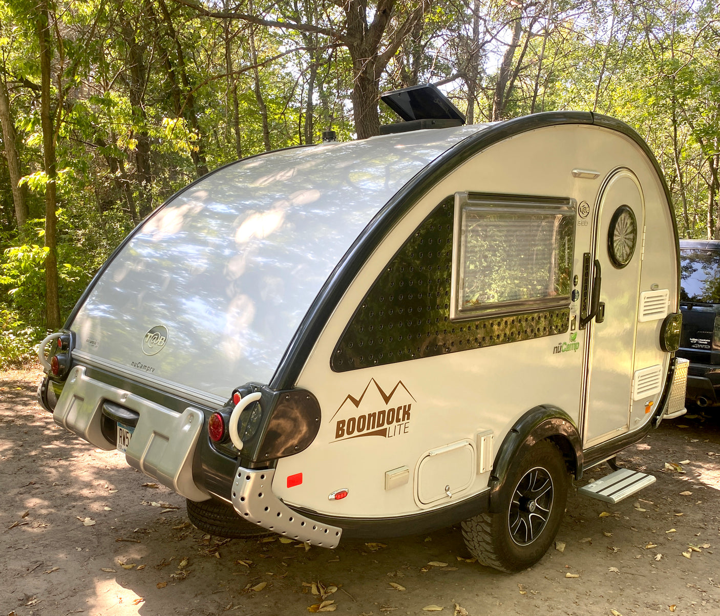 An RV rental, a sleek nuCamp T@B teardrop camper, sits in a sunny, wooded campsite, ready for outdoor adventures.