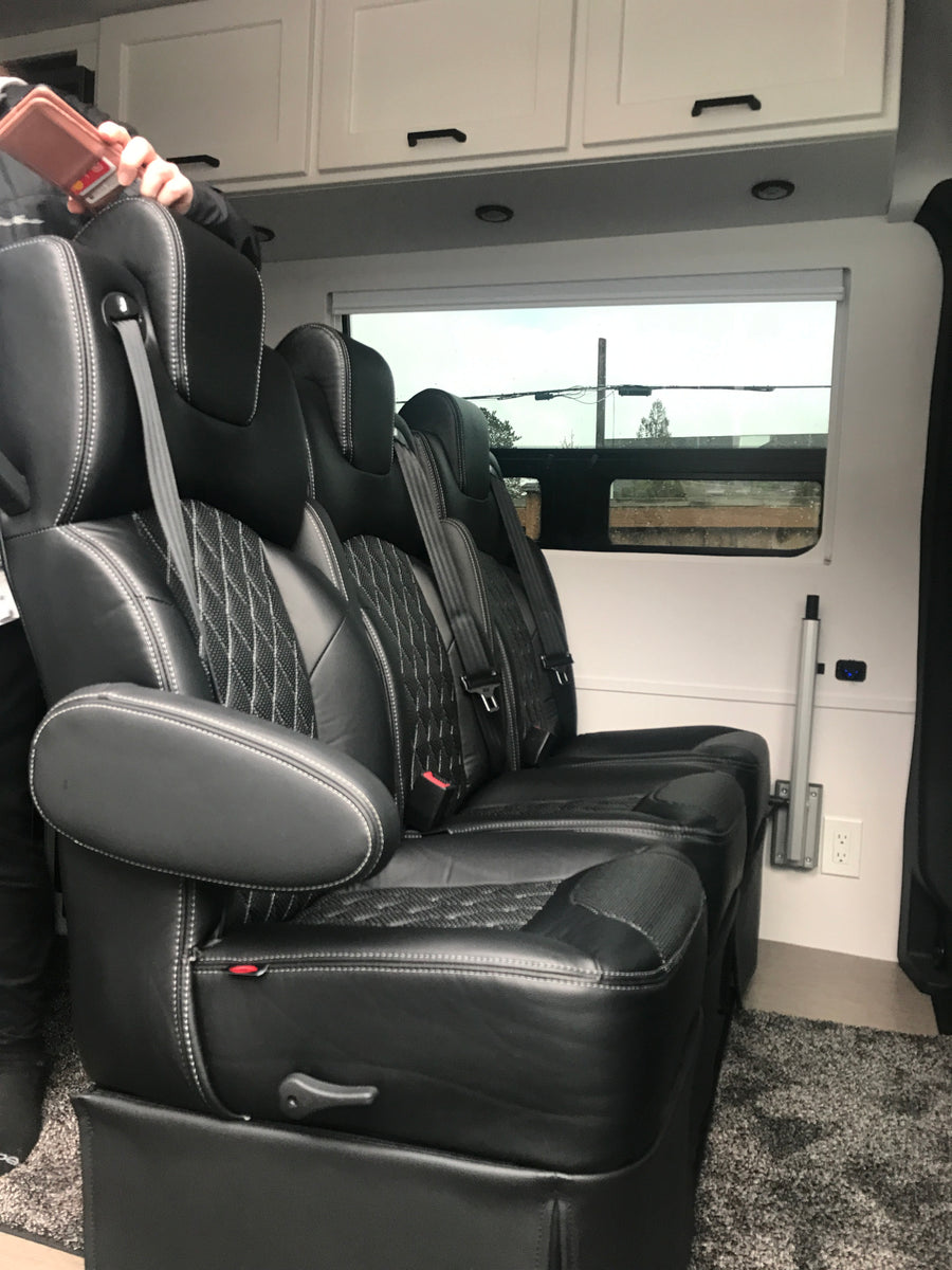 Luxury RV rental interior featuring plush black leather captain's chairs with white stitching, overhead cabinets, and a window view.