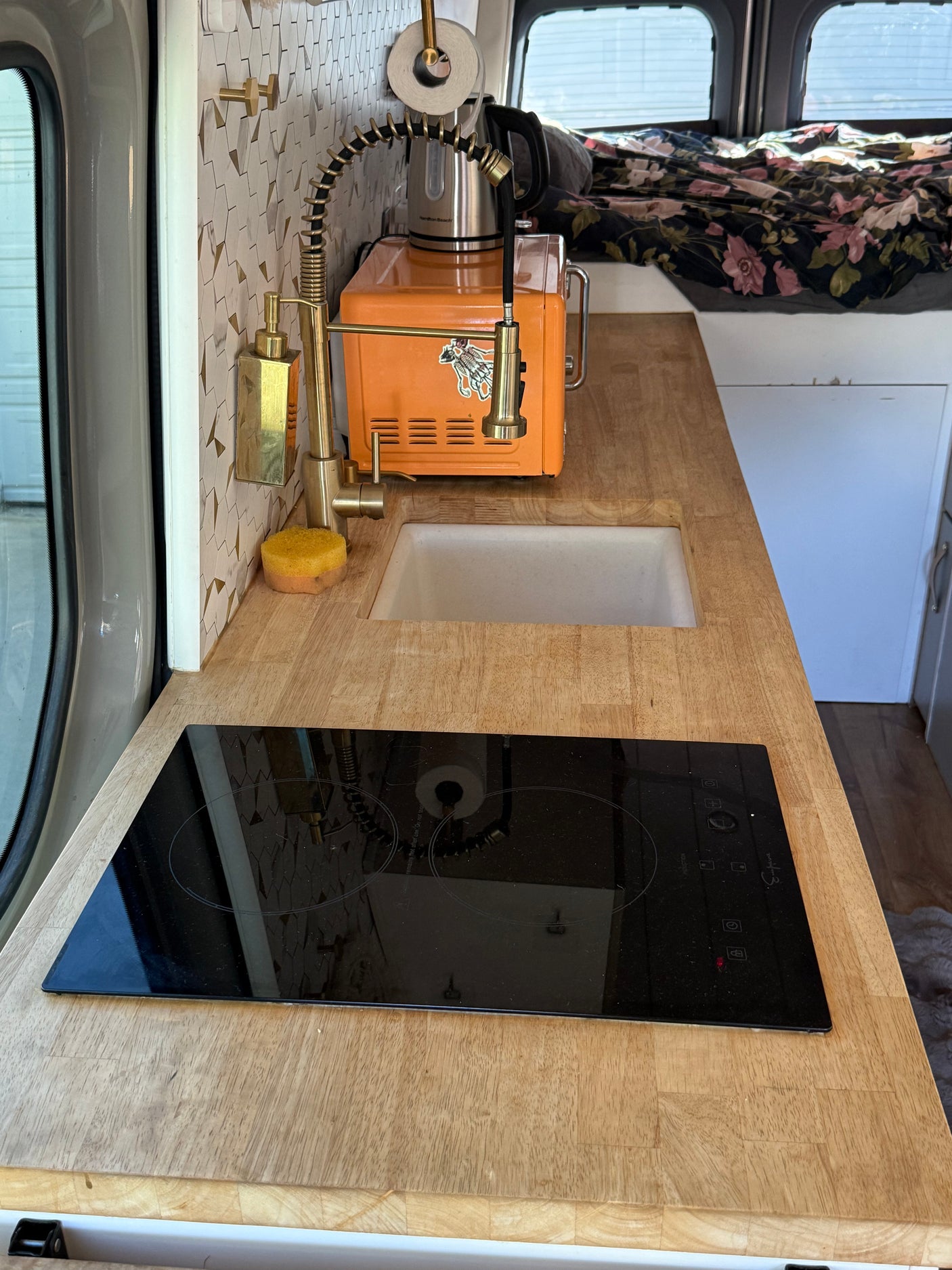 Inside an RV rental: a stylish compact kitchen with wood countertops, sink, induction cooktop, and gold fixtures for van life.