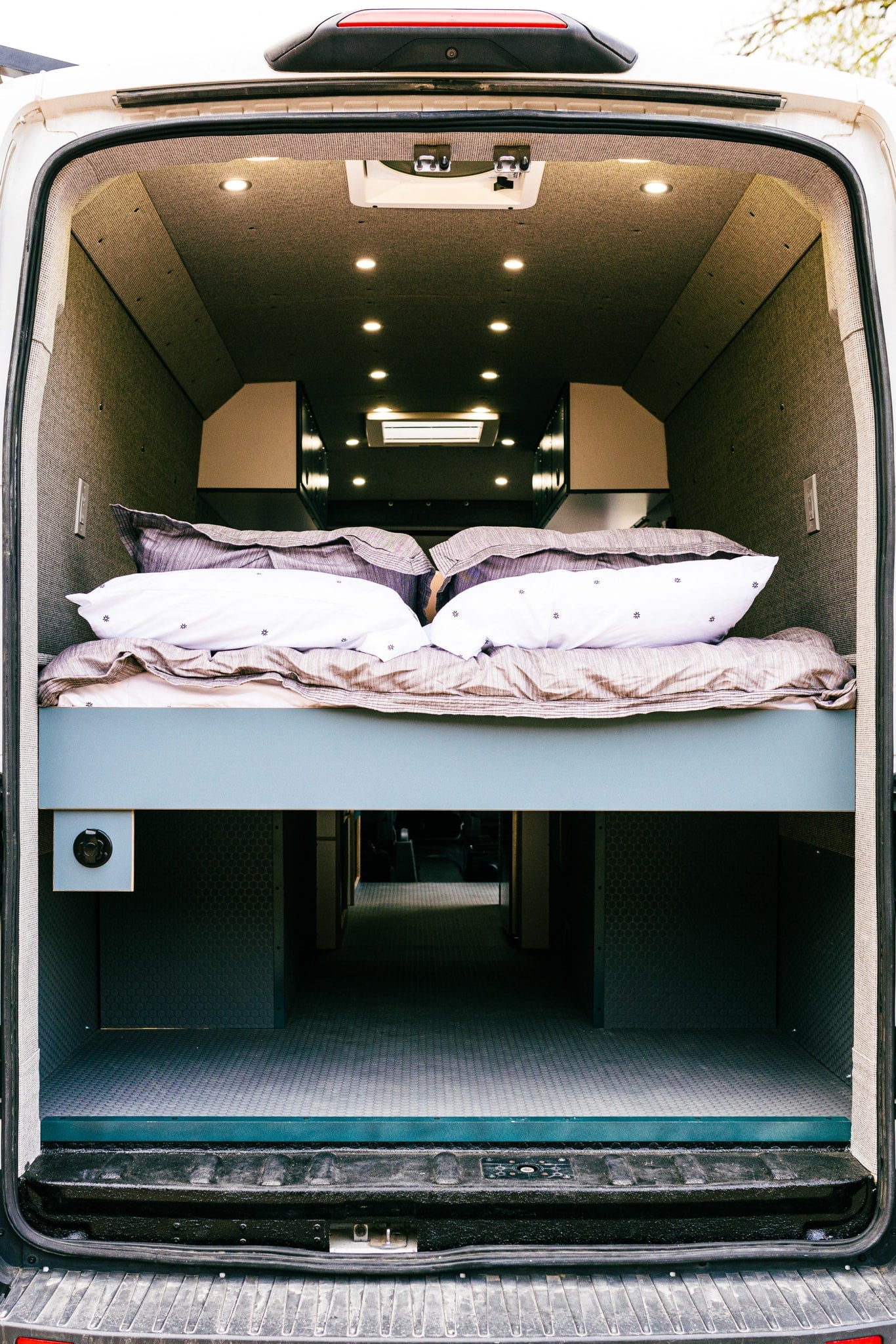 Rear view inside a modern RV rental, featuring a raised bed with pillows and sheets, and a large storage area below. — photo 3