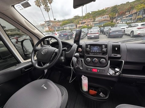 Dashboard & driver's seat of an rv rental campervan. View out to a busy parking lot, coastal houses, and palm trees.