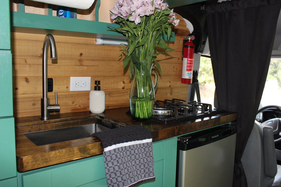 This RV rental's functional kitchen features a modern sink, stovetop, mini-fridge, wood walls, and vibrant teal cabinets.