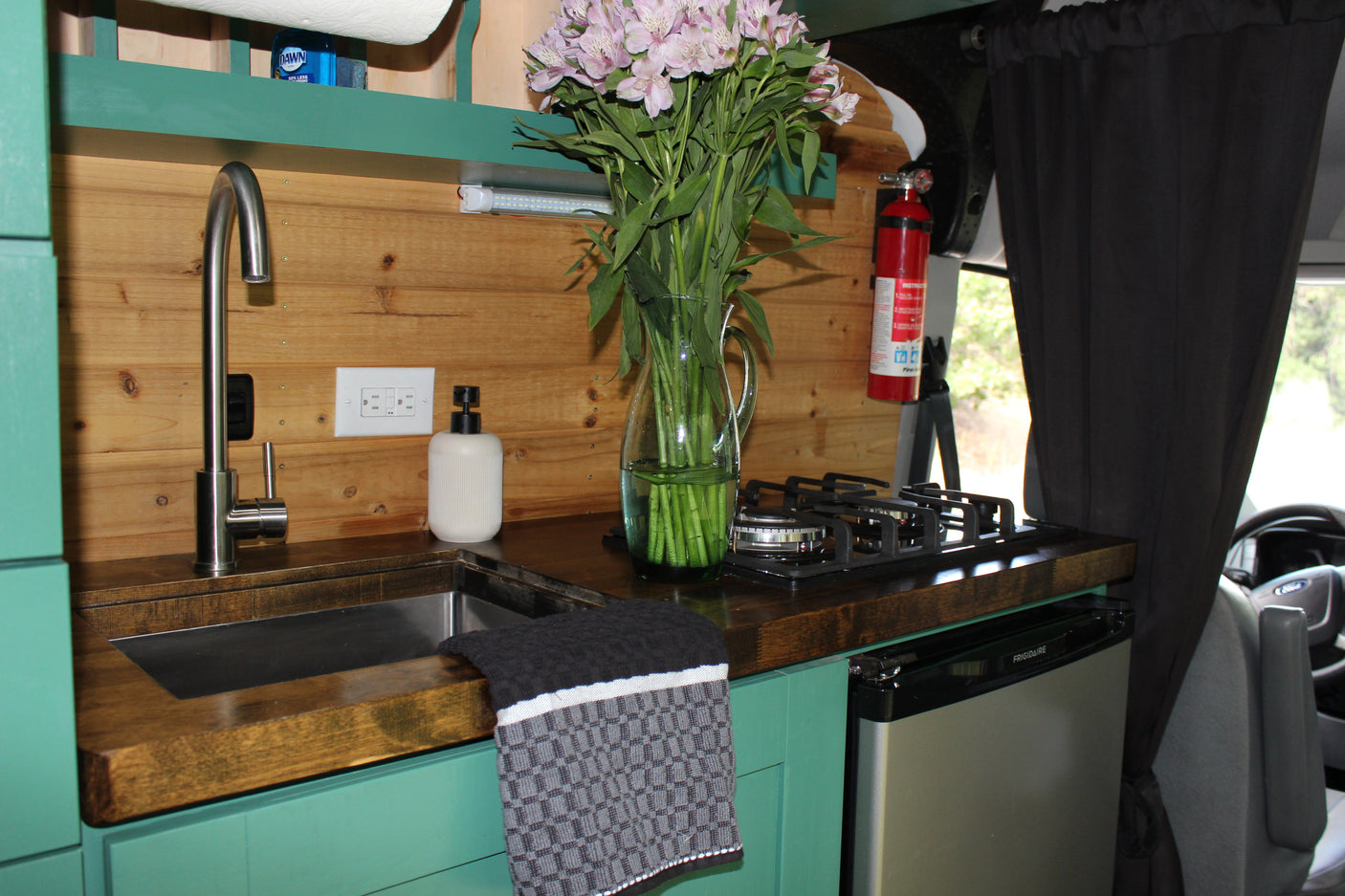This RV rental's functional kitchen features a modern sink, stovetop, mini-fridge, wood walls, and vibrant teal cabinets.
