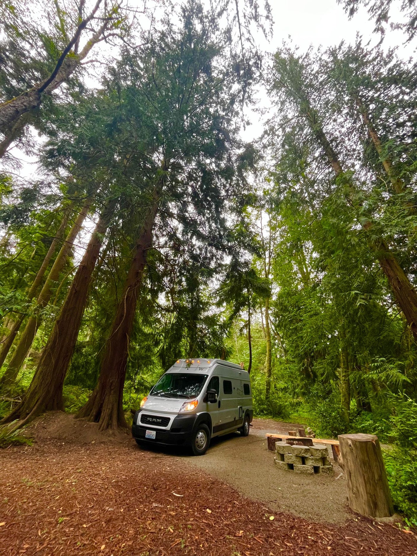 Explore Washington's nature! This RV rental campervan is set up at a serene forest campsite with a stone fire pit.