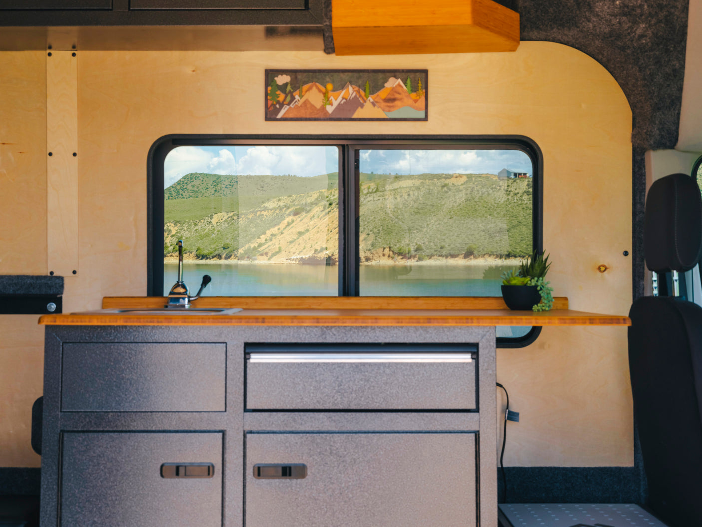 Inside an RV rental: a modern camper kitchen with sink, wooden countertop, and panoramic windows framing a serene lake and mountains.