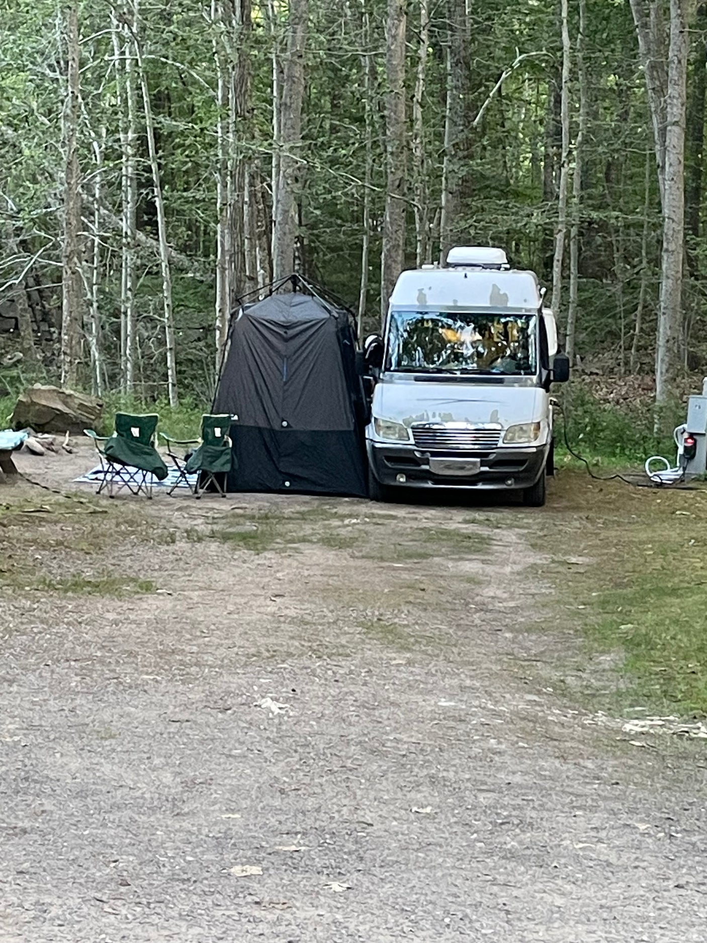 An RV rental camper van at a wooded campsite, with a privacy tent and camping chairs. Ready for outdoor adventure and relaxation.