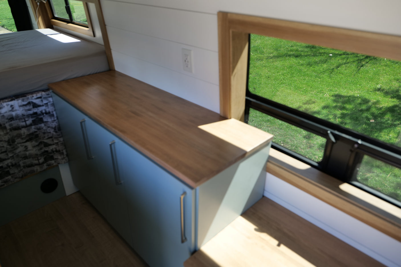 Bright RV rental interior featuring a bed, wood countertop, blue cabinets, and a window with a lush green view, ideal for travel.