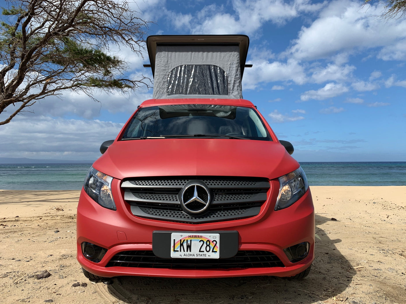 Red Mercedes-Benz campervan RV rental on a Hawaii beach with its pop-top extended. Clear blue sky and ocean in background.